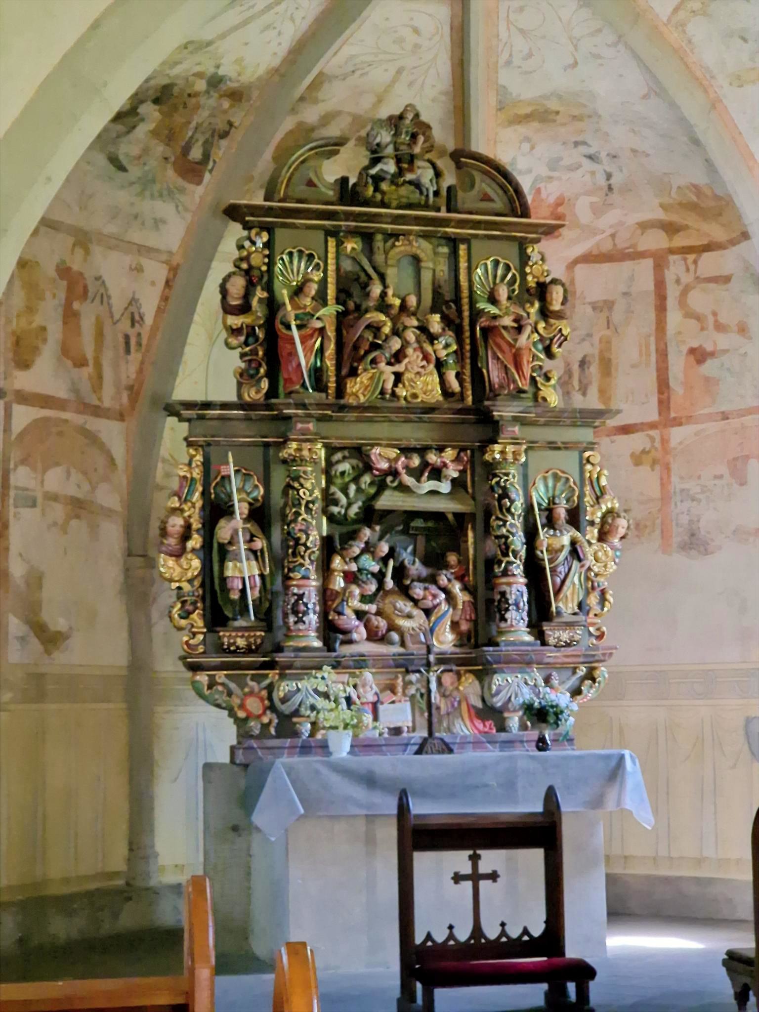 Sankt Jost_Altar_Andachten Stellen Sie sich vor, Sie stehen oder sitzen in einer kleinen Kapelle. Vor Ihnen befindet sich ein reich verzierter, farbig gefasster Altar aus Holz. Der Altar ist mehrstöckig aufgebaut und wirkt wie ein kleines, kunstvolles Haus mit Säulen, Figuren und Nischen. Die Farben sind warm: Gold, Rot, Grün und dunkles Holz prägen das Gesamtbild. Im Zentrum des Altars, auf Augenhöhe, sehen Sie eine Darstellung der Geburt Jesu: Ein kleines Kind liegt in der Mitte, umgeben von mehreren Figuren. Einige Figuren knien, andere stehen. Alle sind dicht um die Mitte gruppiert, sodass ein Gefühl von Nähe und Sammlung entsteht. Darüber, auf einer erhöhten Ebene, befinden sich weitere Figuren, die Szenen aus dem Leben Jesu oder aus der biblischen Geschichte zeigen. Diese Figuren sind kleiner, aber ebenfalls reich gestaltet. Auch hier stehen mehrere Gestalten beieinander, eingerahmt von Säulen und Bögen. Seitlich stehen größere Figuren in Nischen. Jede Figur ist einzeln hervorgehoben, mit Gewändern in kräftigen Farben und goldenen Details. Unterhalb der Hauptszene gibt es eine weitere kleine Darstellung, ebenfalls mit mehreren Figuren, die sich um eine zentrale Szene gruppieren. Vor dem Altar steht ein schlichter, heller Altartisch mit einem weißen Tuch. Darauf befinden sich frische Blumen. Im Hintergrund erkennt man die Wände der Kapelle mit zarten, teilweise verblassten Malereien. Die Decke ist gewölbt und trägt ebenfalls helle, zurückhaltende Bildmotive. Insgesamt entsteht ein Eindruck von Stille, Konzentration und Tiefe: Der kunstreiche Altar lädt dazu ein, den Blick immer wieder zur Mitte zu führen – dorthin, wo das Wesentliche liegt.